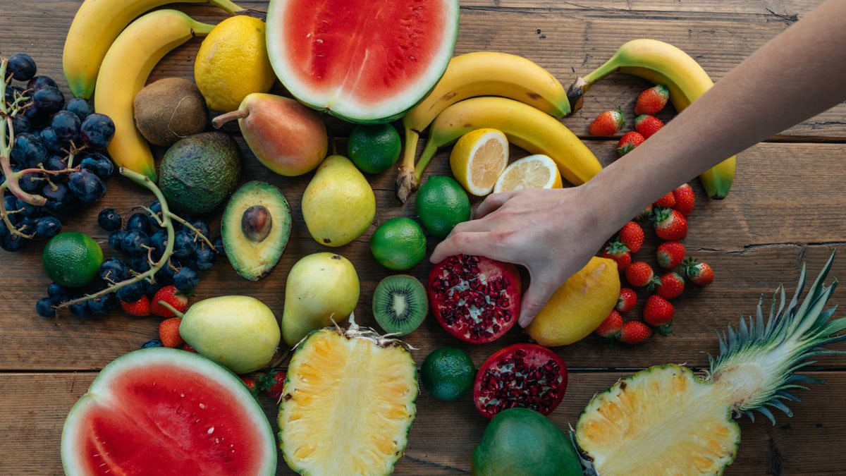 Colorful variety of fresh fruits arranged on a wooden surface with a hand selecting a pomegranate.